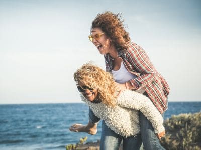 Outside by the sea one lady is giving another lady a piggy back - both are laughing symbolising how a sense of humour keeps you young at heart