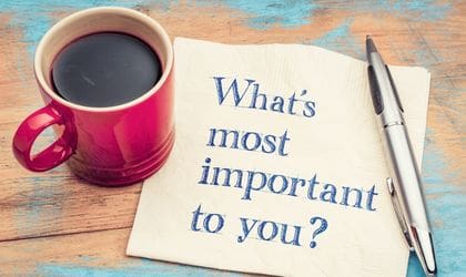 There is a close up of a wooden table with a red mug with black coffee in it, next to it is a napkin with the words printed on of it of 'What's most important to you?' and a pen next to this. It is being used to represent the importance of values